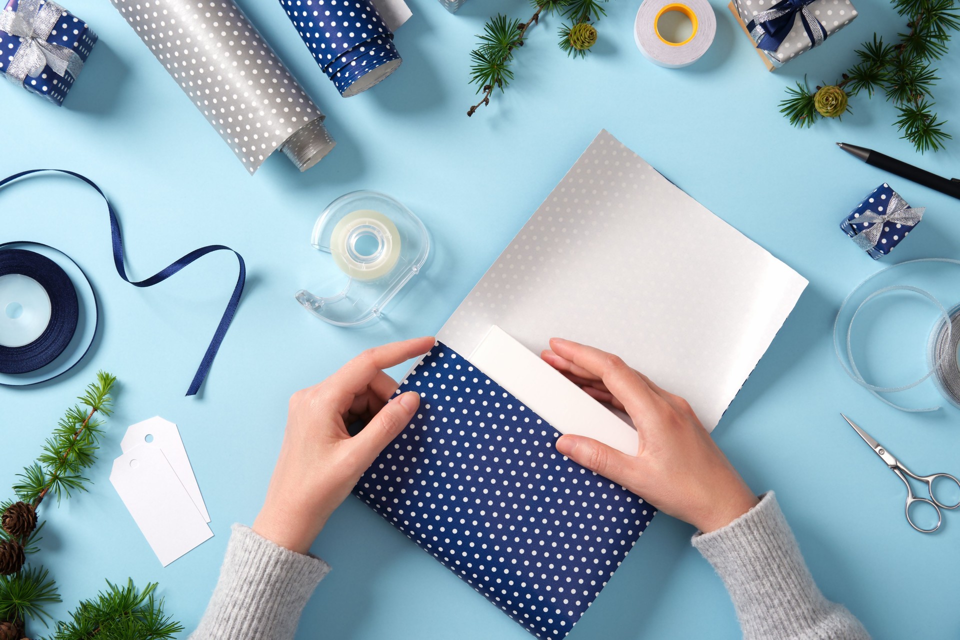 Hands wrap the box in wrapping paper. Around it are ribbon, scissors, tape, spruce twigs, and small wrapped gifts. Preparing for the holidays.